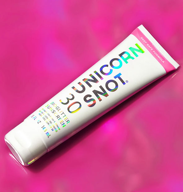 Tube of Unicorn Snot BioGlitter Sunscreen with pink stripe and holographic lettering on a bright pink surface