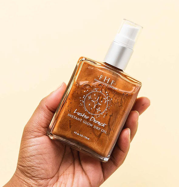 Model's hand holds a square glass bottle of FarmHouse Fresh Lustre Drench Instant Glow Dry Oil which has a shimmery bronze appearance
