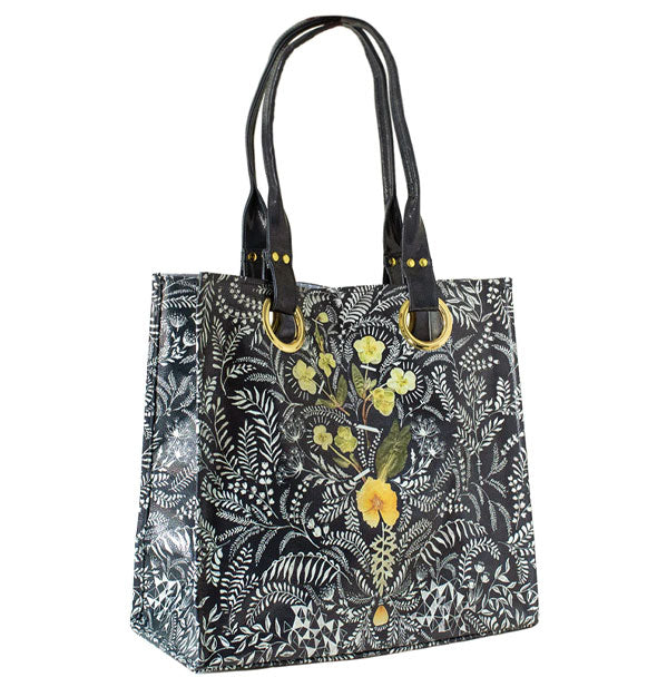Black tote bag with all-over intricate white floral brushstrokes and central yellow pressed flower design has two shoulder straps accented by gold hardware