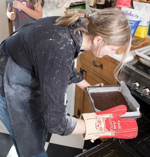 A flour-spattered woman wearing the Made From Scratch oven mitt puts a brownie pan into an oven