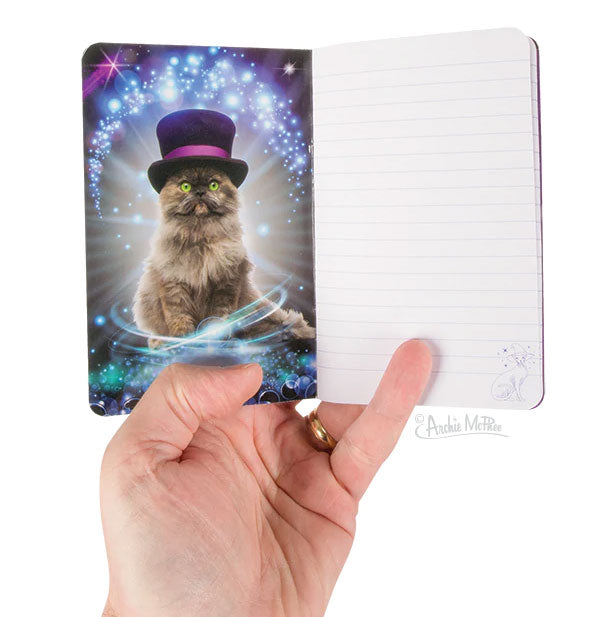A hand holds open a notebook with illustration of a long-haired cat in a top hat surrounded by stars and light flares alongside a ruled page