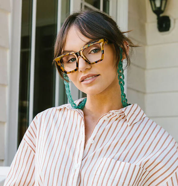 Model wears a square pair of oversized reading glasses with a large green chain attached to the arms