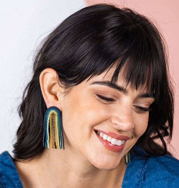 Smiling model wears a pair of beaded rainbow fringe earrings in blue and gold tones