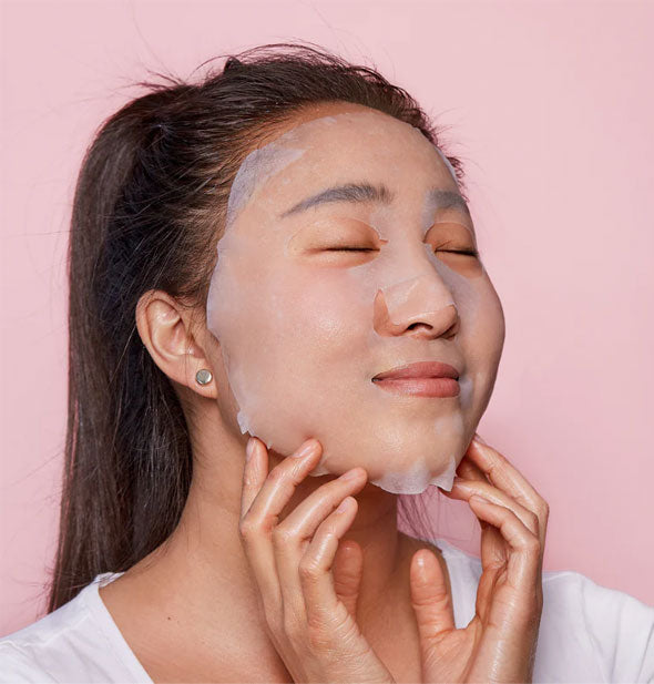 Smiling model lightly touches a sheet mask applied to face