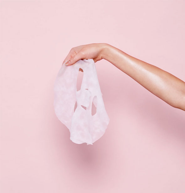 Model's outstretched hand holds a sheet mask in front of a pink background