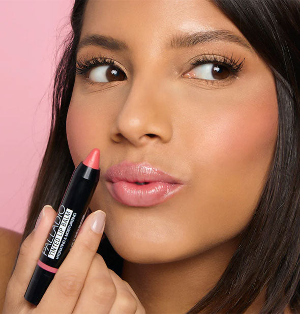 Model puckers lips to apply Palladio Tinted Lip Balm in a pink shade
