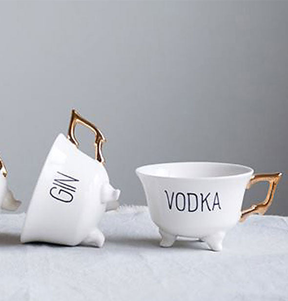 White footed teacups with gold handles say "Gin" and "Vodka" respectively