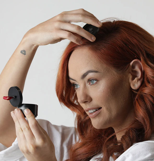 Model looks into the mirror in the lid of a compact of Style Edit Root Touch-Up Binding Powder while working product into red hair