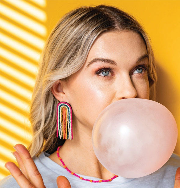 Model blowing a bubble wears a pair of rainbow beaded fringe earrings