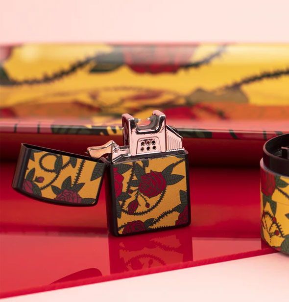 Flip-top lighter with silver hardware inside features a red thorny rose print on a yellow background; lighter rests on a red surface with matching accoutrements