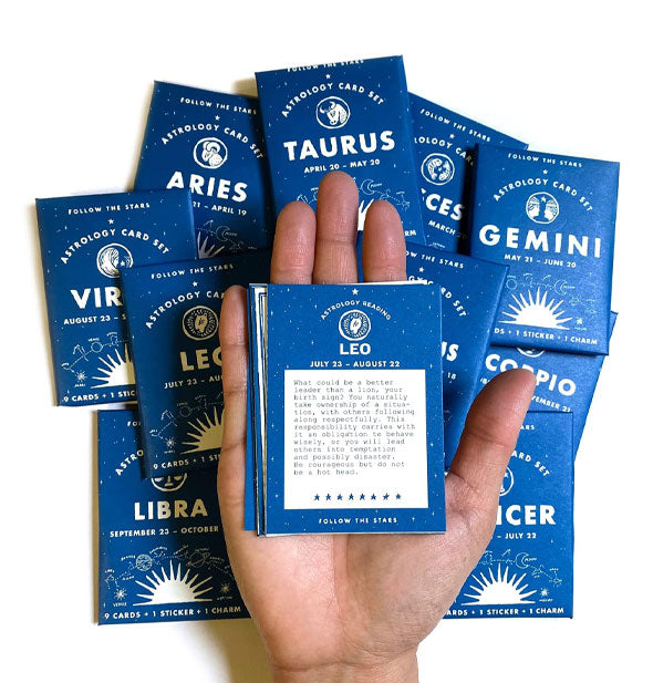 Model's hand holds a pack of Leo Astrology Cards above an assortment of Astrology Card Sets representing the other signs of the zodiac