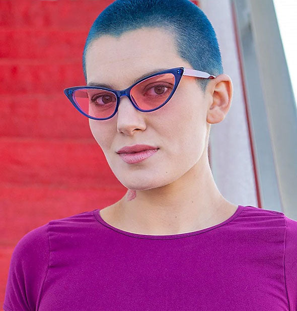 Model wears a pair of blue cat-eye sunglasses with rose-colored lenses