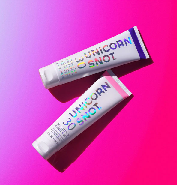 Two tubes of Unicorn Snot BioGlitter Sunscreen, one with a purple stripe and one with a pink stripe, on a purple-to-pink ombre surface