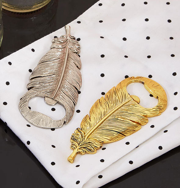 Two feather shaped bottle openers in silver and gold finishes on and black and white polka dotted cloth