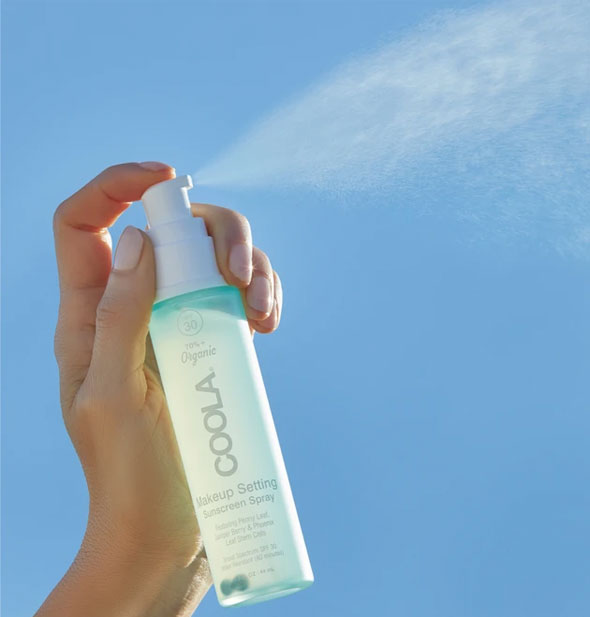 A model's hand holds and sprays a bottle of COOLA Makeup Setting Sunscreen Spray in front of a blue sky