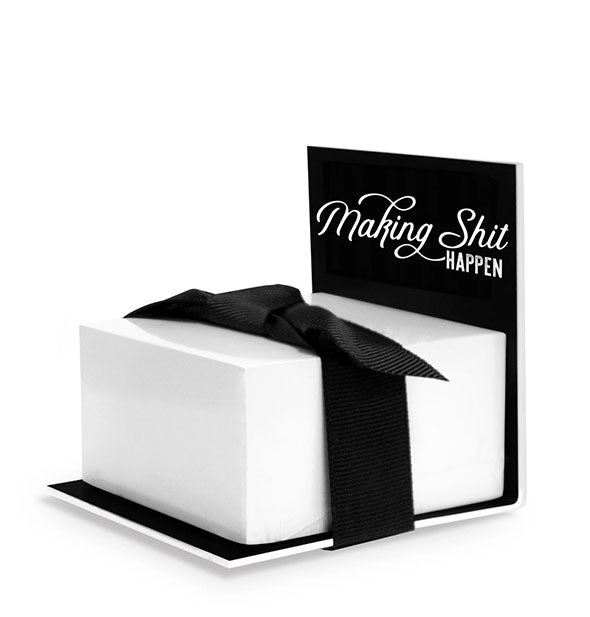 A stack of white sticky notes wrapped in a black bow sits on an L-shaped display printed with the words, "Making Shit Happen" in white lettering of alternating type styles