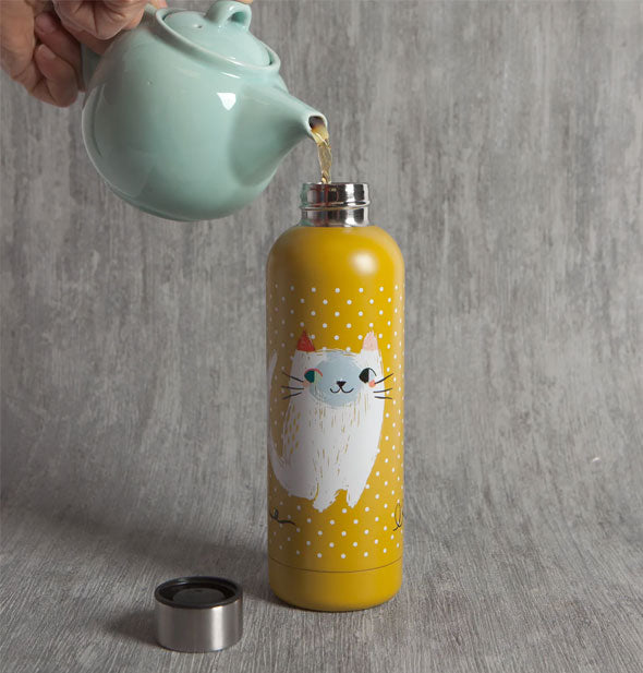 Model's hand pours tea from an aqua-colored teapot into a stainless steel-topped yellow water bottle with white cat artwork