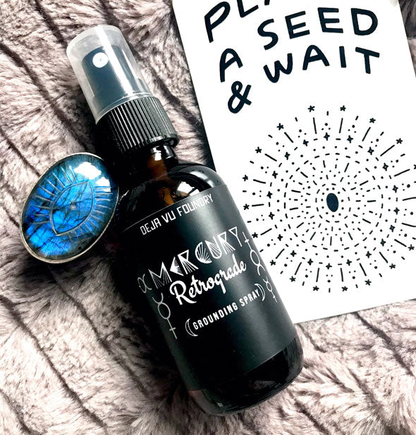 Amber bottle of Mercury Retrograde Grounding Spray with black label rests on a plush cushion with evil eye crystal and tarot card