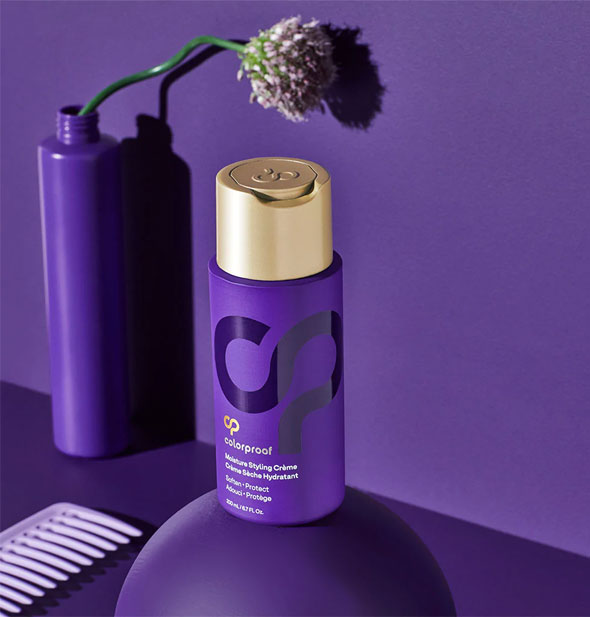 Bottle of ColorProof Moisture Styling Crème staged with a flower in a slender purple bottle, light purple comb, and purple sphere on a purple background