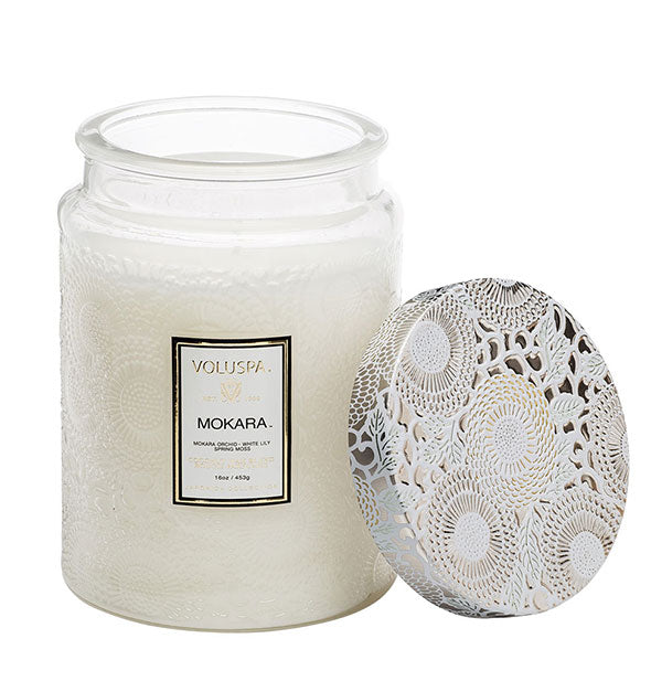 White embossed glass Mokara Voluspa candle jar with white and gold metallic floral lid set to the side