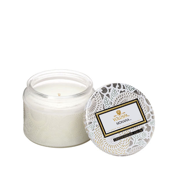 White embossed glass Mokara Volspa jar candle with white, silver, and gold metallic floral lid set to the side