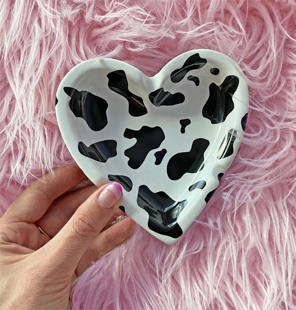 Model's hand holds a black and white cow print heart-shaped ashtray against a pink fur backdrop