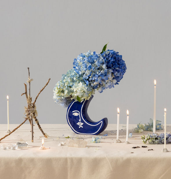 Blue crescent moon vase filled with blue hydrangeas is staged on a tabletop with taper candles, flower petals, and freestanding twig sculpture