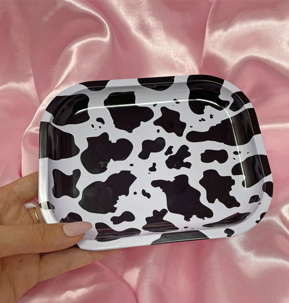 Model's hand holds a rectangular tray with rounded corners and black and white cowhide print in front of a pink satin background