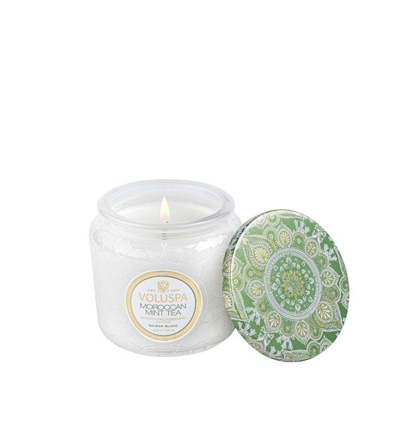 A lit Voluspa Moroccan Mint Tea candle in frosted embossed glass jar with decorative green tin lid alongside