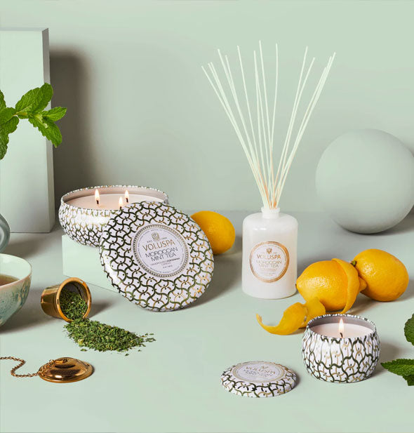 Voluspa tin candles and reed diffuser on a green background are staged with herbs and citrus