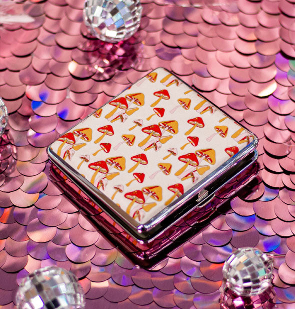 Square cigarette case with  a silver rim and print of red and orange mushrooms on a white background rests on a pink sequined surface with mini disco balls