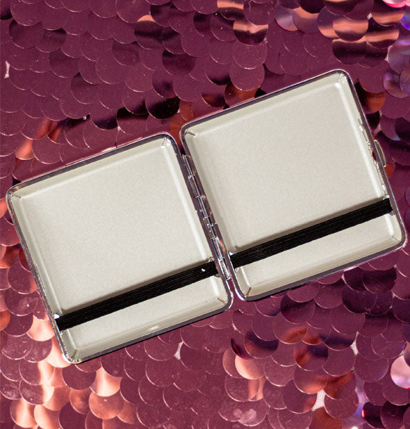 Opened cigarette case with white interior and black elastic bands to secure contents rests on a pink sequined surface