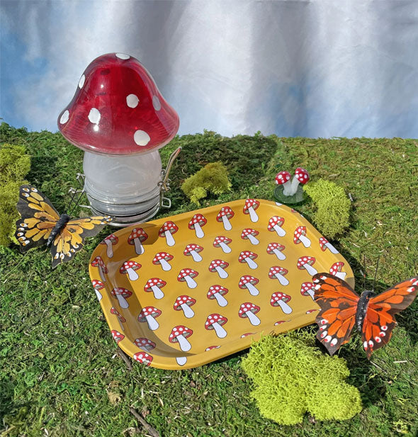 Rectangular yellow tray with rounded corners and red and white mushrooms print rests on a grassy surface with butterflies, moss, and a larger glass mushroom