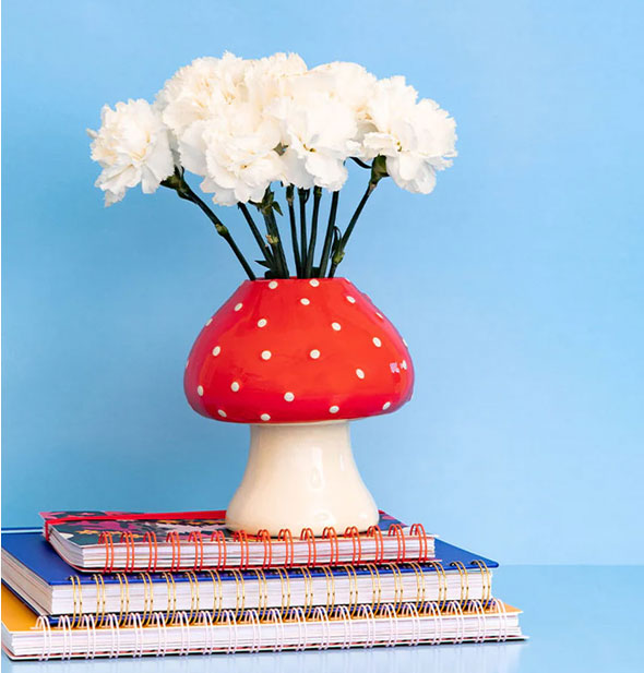 Mushroom vase is staged with white flowers and is placed on top of a stack of three spiral-bound notebooks against a blue background