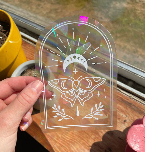 Model's hand holds a clear sticker with white moth, celestial, and botanical outline designs