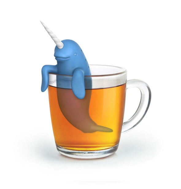 A blue narwhal tea diffuser with white horn rests on the edge of a clear glass mug filled with tea