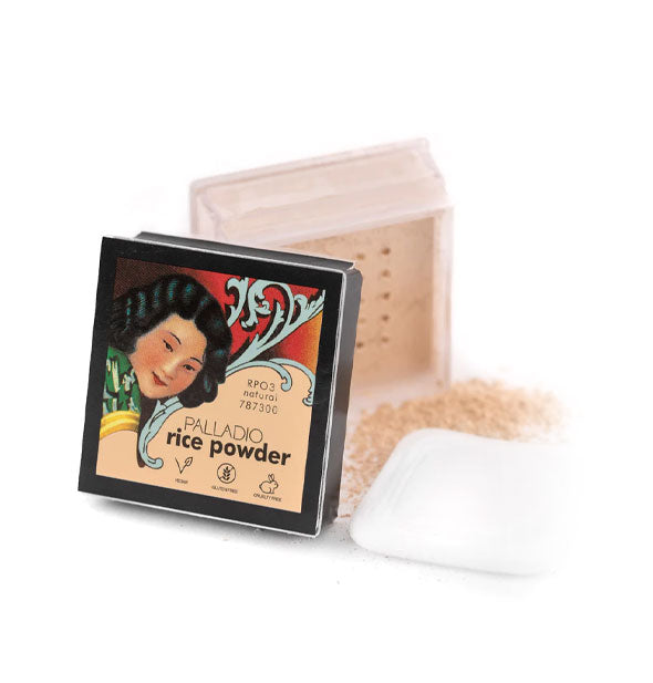 Opened square compact of Palladio Rice Powder with decorative lid placed in the foreground of some scattered product and white puff applicator
