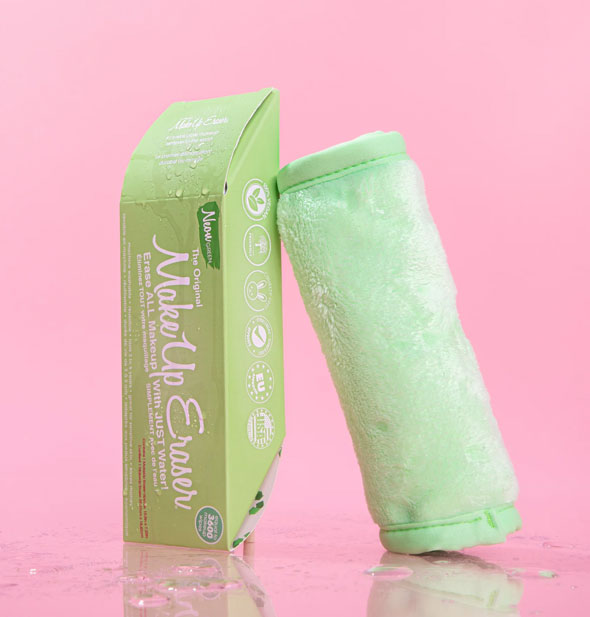 Rolled up Neon Green MakeUp Eraser leans against its box on a pink surface sprinkled with water