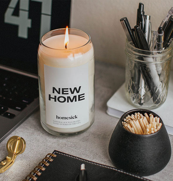 A lit New Home candle sits on a desktop with office supplies and matches