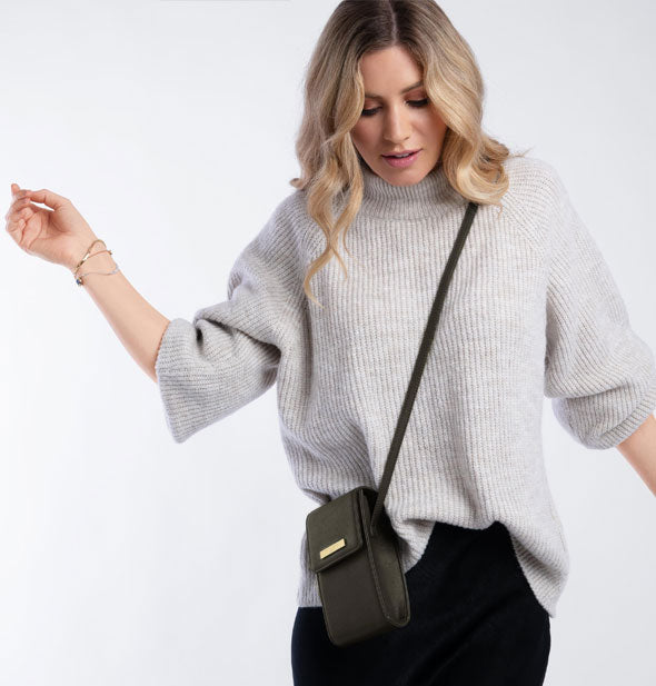 Model with arms outstretched wears a dark green crossbody bag across her shoulder