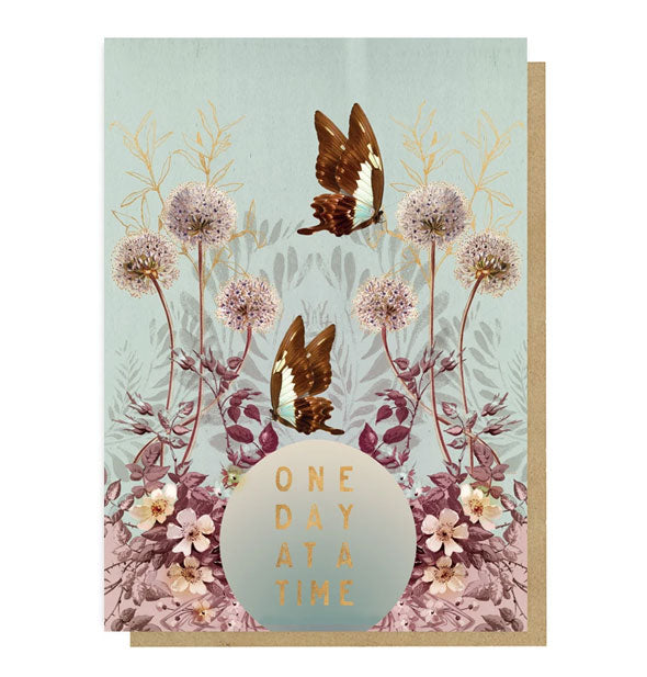 Dusty blue ombre greeting card with kraft envelope behind features illustrated wildflowers and butterflies above the words, "One Day at a Time" in metallic gold foil lettering