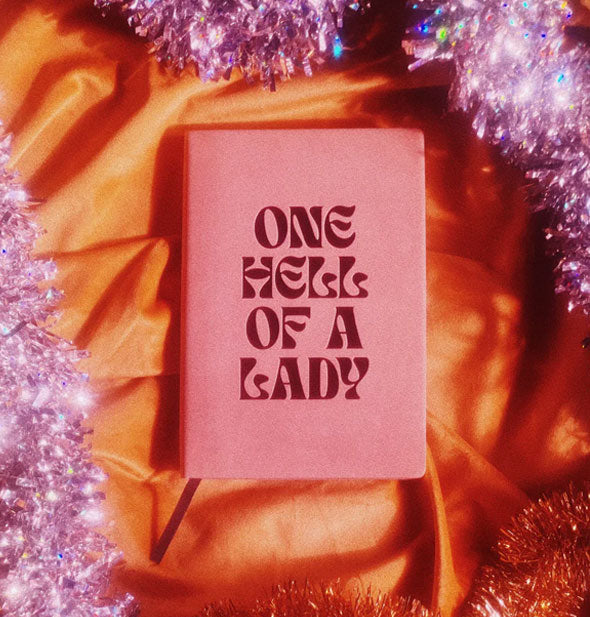 One Hell of a Lady journal rests on a piece of cloth surrounded by purple tinsel