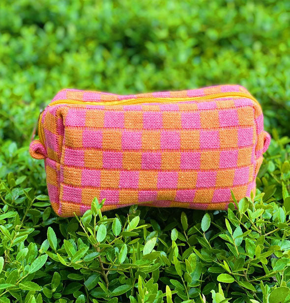Orange and pink checker print knit makeup bag rests on a green hedge