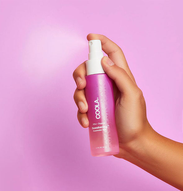 A model's hand holds and sprays from a bottle of Coola Rewakening Rosewater Mist Face Spray