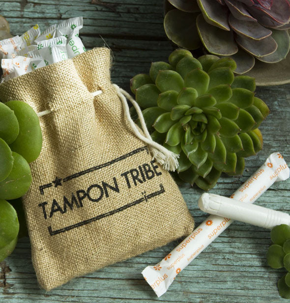 Burlap cinch bag stamped with "Tampon Tribe" graphic holds several assorted tampons and is staged with an opened tampon along with succulent plants on a wooden surface