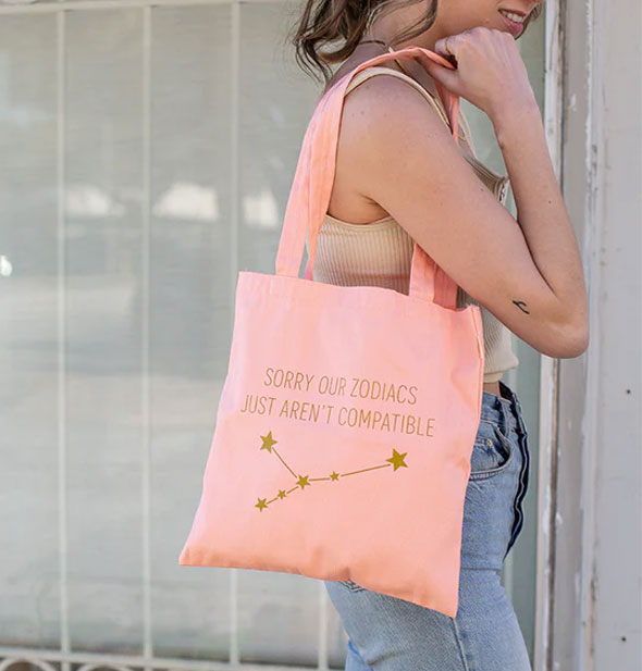 Smiling model wears a pink tote bag over shoulder that says, "Sorry our zodiacs just aren't compatible" above a gold star constellation graphic