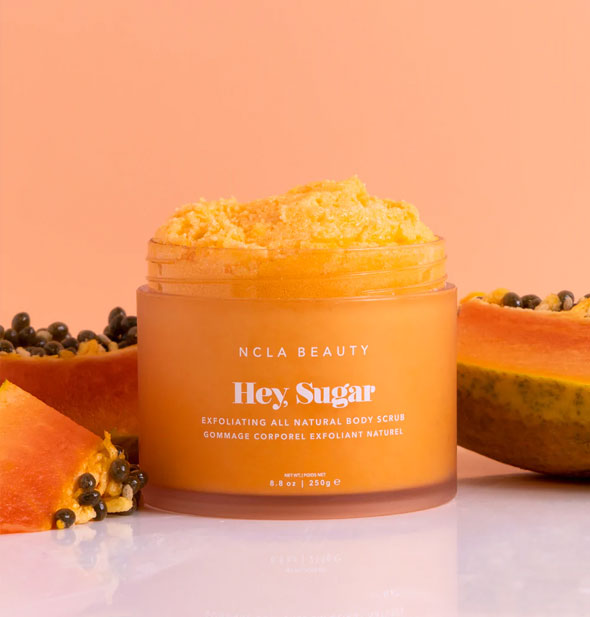 Jar of orange Hey Sugar Exfoliating All Natural Body Scrub by NCLA Beauty is staged with slices of papaya