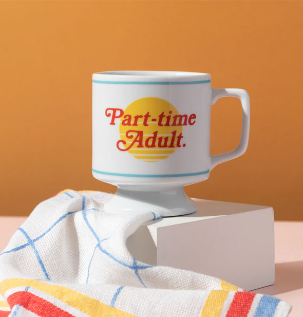 Part-time Adult coffee mug on a block platform with white, blue, red, and yellow dish towel draped in front