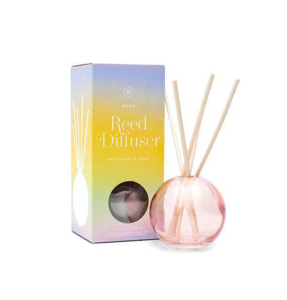 Round pink glass reed diffuser with five wooden reeds sticking out of it sits next to box packaging