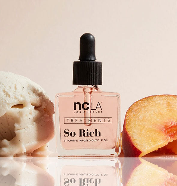 Square glass bottle of NCLA Treatments So Rich cuticle oil with black rubber dropper cap is staged with vanilla ice cream and peach slices